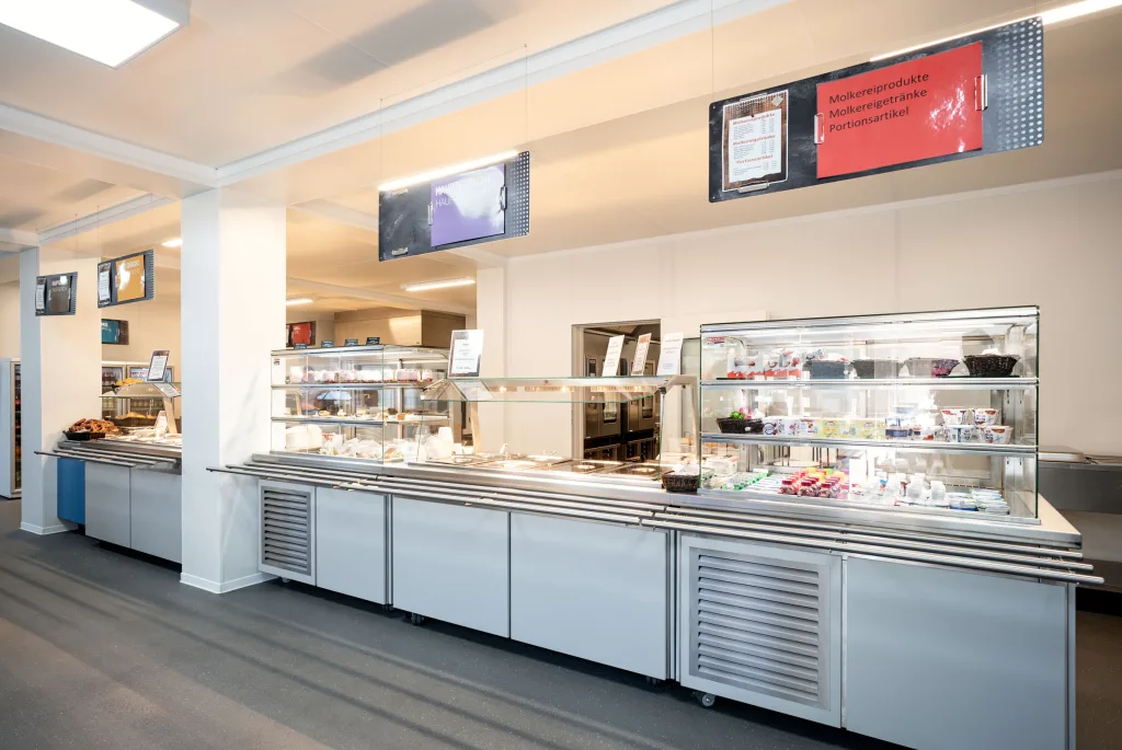 Speisenausgabe der Interimskantine mit Kühltheken, Vitrinen und digitalen Menütafeln.
