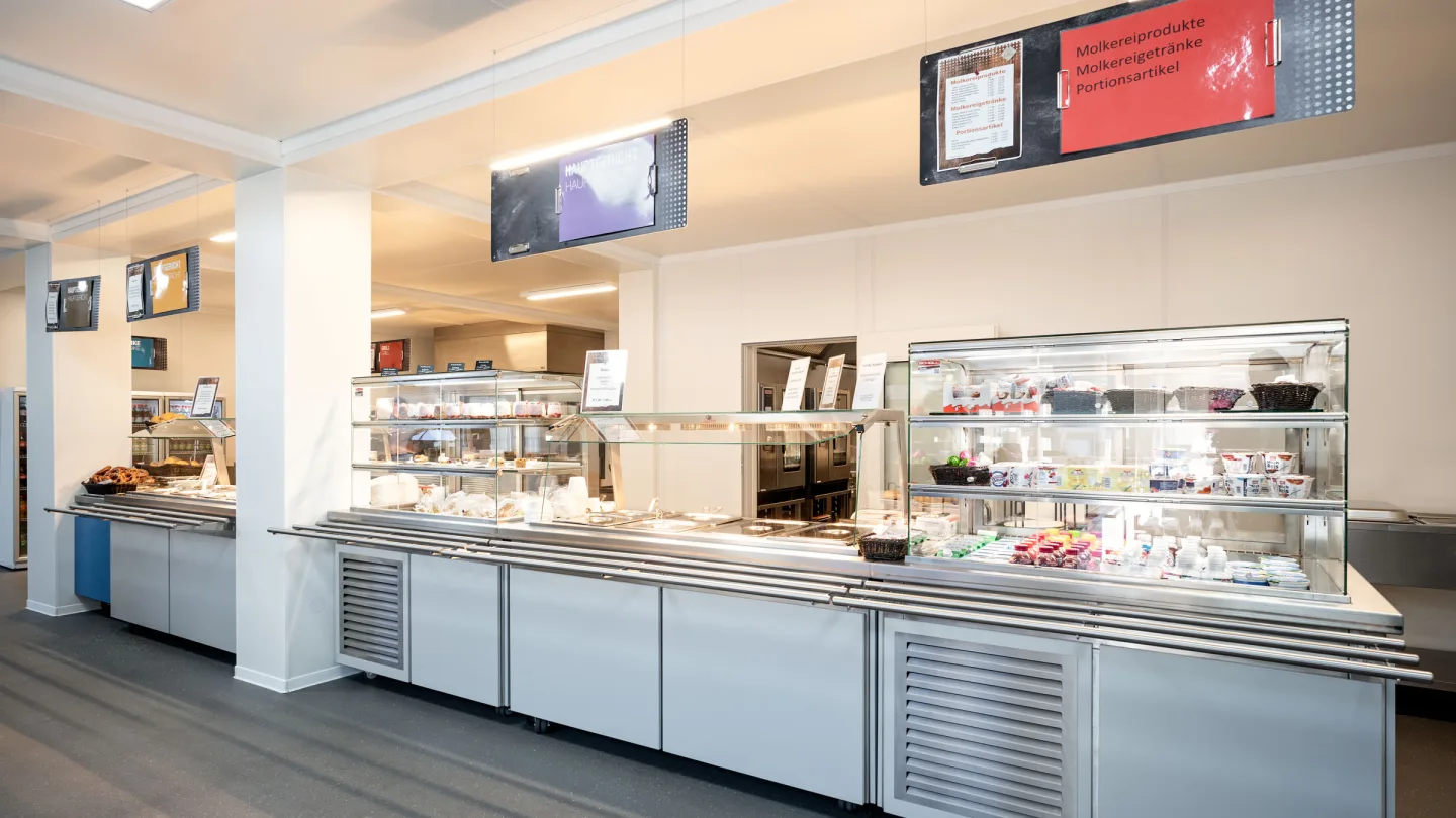 Speisenausgabe der Interimskantine mit Kühltheken, Vitrinen und digitalen Menütafeln.